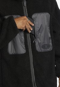 Svart fleecejacka med släta grå nylonlappar, dragkedjefickor och logotypdetaljer. Den mjuka texturen och den avslappnade passformen är framträdande egenskaper.