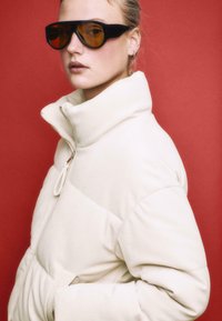 Puffjacke aus cremefarbig geripptem Stoff mit hohem Kragen und Reißverschluss. Große schwarze Sonnenbrille mit bernsteinfarbenen Gläsern. Roter Hintergrund.