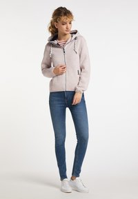 Felpa rosa chiaro con coulisse, chiusura a zip e tasche laterali, abbinata a jeans skinny blu e sneakers bianche. Texture liscia.