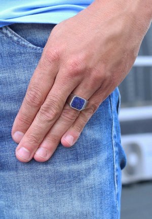 Silberring mit einem quadratischen blauen Stein, der an einer Hand getragen wird, die auf Denim-Stoff ruht. Die Hautstruktur ist um die Finger sichtbar.
