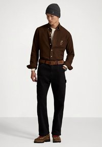 Καφέ πουκάμισο με κουμπιά και λογότυπο στην τσέπη, μαύρο παντελόνι, καφέ ζώνη με υφή και καφέ ορειβατικά μποτάκια με κόκκινες κορδόνια. Γκρι σκουφί.