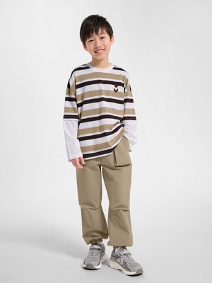 Smilende dreng, der står iført beige bukser, grå sneakers og en langærmet stribet skjorte med beige, sort og hvide horisontale striber.