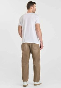Un homme portant une chemise blanche à manches courtes, un pantalon marron clair et des baskets blanches se tient de dos devant un fond clair.