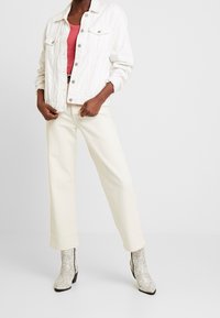 Vit denimjacka över en rosa topp, i kombination med krämfärgade vida byxor och grå ormskinnsankelstövlar, vilket visar en modern avslappnad stil.