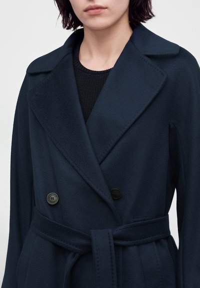 Femme portant un manteau ceinturé bleu marine foncé avec de grands revers et deux boutons visibles sur un top en maille noir.