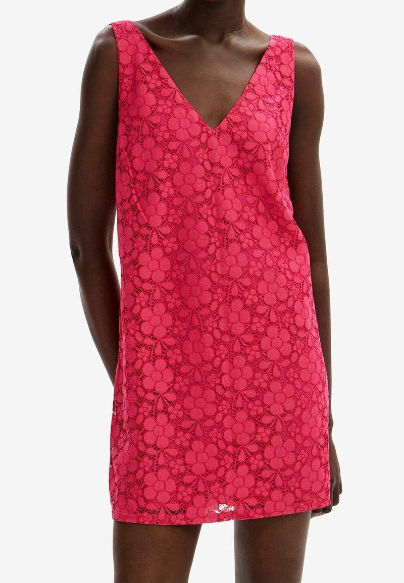 Modelka v bezramínkových růžových květinových krajkových šatech s výstřihem do V, které zdůrazňují složitý krajkový vzor a obepnutou siluetu.