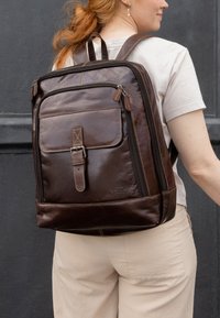 Brown leather backpack featuring a flat front pocket, adjustable straps, and zipper details. Stitched accents and a subtle logo at the base.