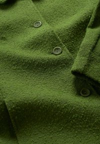 Grön texturerad tyg med en fluffig yta, med två cirkulära knappar. Materialet verkar mjukt och något upphöjt.