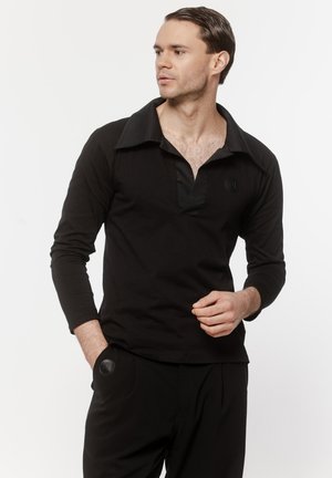 Mężczyzna w czarnej koszuli z długim rękawem o szerokim kołnierzu i czarnych spodniach, patrzący w bok, z jedną ręką w kieszeni.