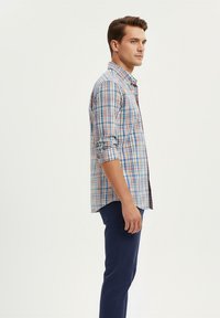 Hombre joven de pie de lado, vistiendo una camisa de cuadros de manga larga con las mangas arremangadas y pantalones azul oscuro, contra un fondo plano.