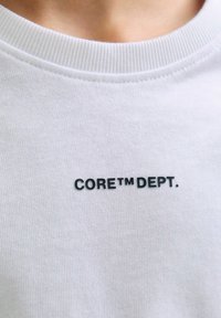 Bílá tričko s kulatým výstřih a černým textem "CORE™ DEPT." umístěným ve středu pod límcem na hrudi.