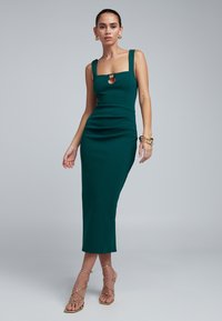 Turquoise midi-jurk met een vierkante halslijn en een open detail. Aansluitend model met textuurstof en zijopeningen, gecombineerd met gouden sandalen met bandjes.