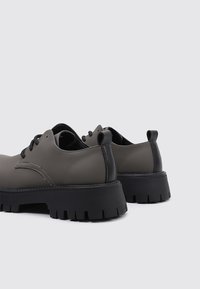 Zapatos de cuero sintético gris con puntera redondeada, suela de goma negra y patrón de borde visible. Presenta cordones negros y acentos de costura.