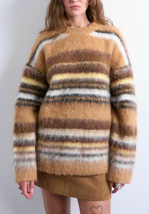 Femme portant un pull rayé et duveteux aux tons marron, jaune, blanc et gris, associé à une mini-jupe en daim marron et des bagues aux deux mains.