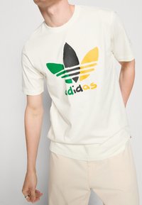 Krémové bavlněné tričko s krátkými rukávy, zdobené vícolor logem Adidas: černé, zelené, žluté a bílé akcenty.