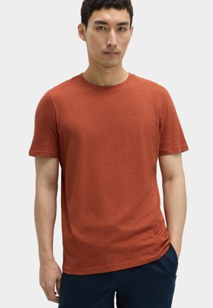 Junger Mann trägt ein schlichtes, rostfarbenes Kurzarmshirt und dunkle Hose und steht vor einem schlichten hellen Hintergrund.