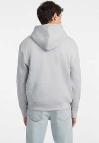 Felpa grigia con cappuccio realizzata in tessuto misto cotone, caratterizzata da un taglio morbido e maniche a coste, abbinata a jeans azzurri chiari.