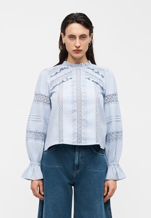 Woman wearing a light blue long-sleeve blouse with lace details and ruffled cuffs, paired with high-waisted blue jeans.