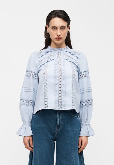 Kvinna som bär en ljusblå blus med lång ärm, spetsdetaljer och volanggardiner, kombinerad med högmidjade blå jeans.