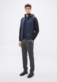 Tumši zils kapucētas pāršūtas vestes virs tumši zila džempera, kopā ar pelēkām biksēm. Piedāvā melnas detaļas uz piedurknes un melnus potīšu zābakus.