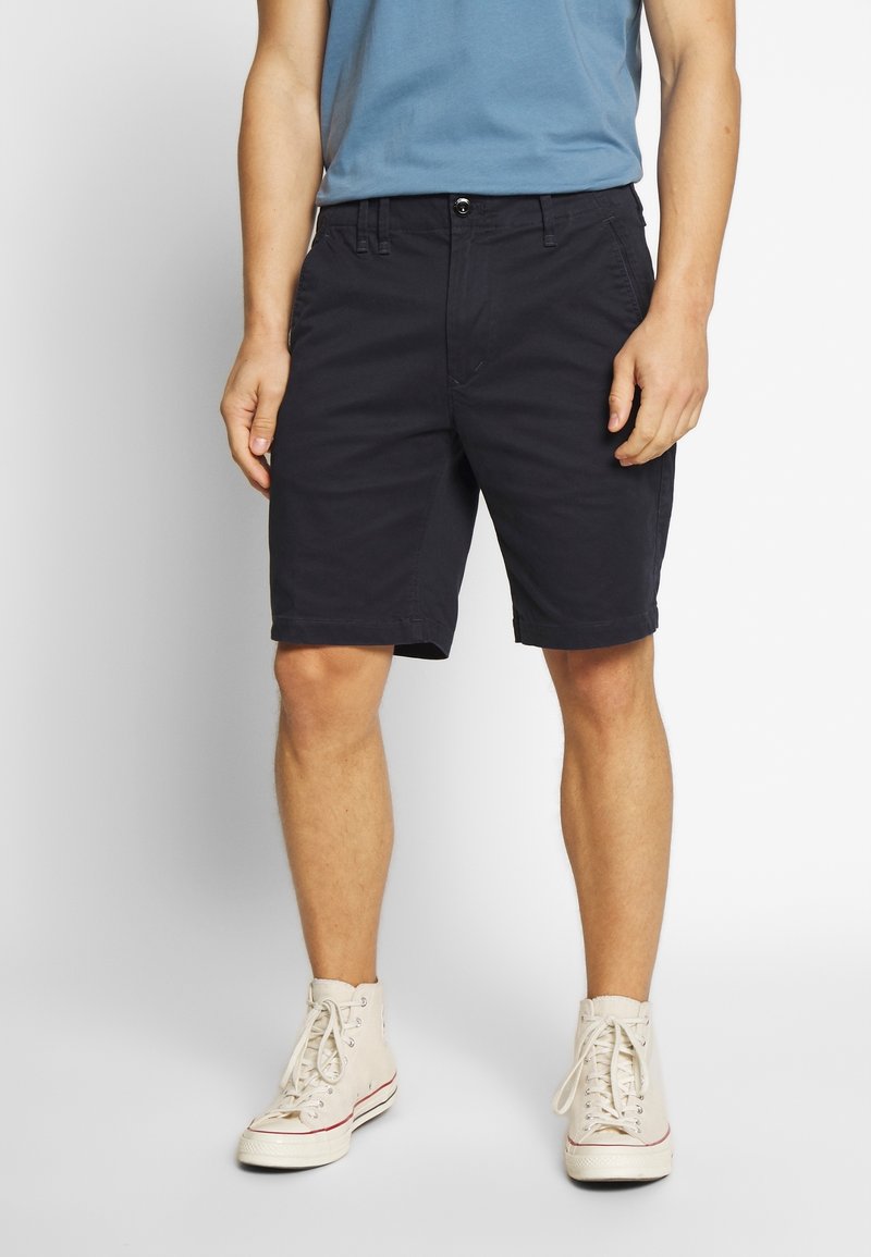 Homme portant un short noir mi-genoux, un t-shirt bleu clair et des baskets montantes blanches, debout devant un fond uni.