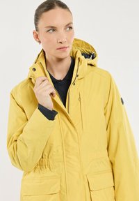 Veste imperméable jaune avec capuche, fermeture éclair à l'avant, taille élastique et poches latérales. Présente une texture lisse et des accents noirs à l'intérieur.