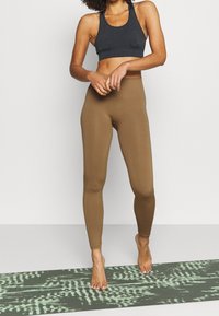 Reggiseno sportivo nero con scollatura a barco e fascia larga, abbinato a leggings marroni aderenti. Il modello è in piedi su un tappetino da yoga verde con texture.