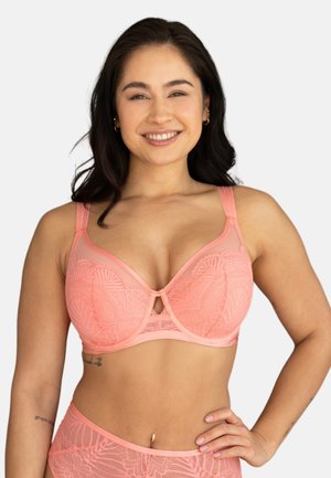 Lächelnde Frau mit dunklem Haar trägt rosa Spitzen-BH und passende, hoch taillierte Unterwäsche, Hand auf der Hüfte, einfarbiger heller Hintergrund.