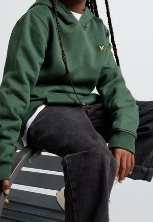 Osoba w zielonej bluzie z kapturem z wyszywanym żółtym emblematem, czarnych spodniach i białych trampkach, siedząca na szarej ławce.