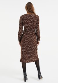 Lång brun klänning med vitt och blått geometriskt mönster, elastisk midja, långa ärmar och längd till mitten av vaden. Svarta ankellånga stövlar synliga.