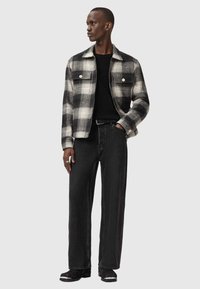 Veste à carreaux noir et blanc, à boutons avec deux poches poitrine, portée avec un pull noir et un pantalon en denim sombre ample. Chaussures noires avec des accents argentés.