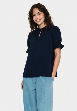 Blouse bleu marine à manches courtes avec un décolleté en forme de trou de serrure et des poignets smockés, associée à un jean large bleu clair. Texture de tissu lisse, coupe décontractée.