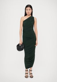 Sukienka na jedno ramię w ciemnozielonym kolorze z teksturowanym marszczeniem, paskami przy kostkach oraz czarną, zakrzywioną torebką. Posiada dopasowaną sylwetkę.