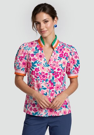 Vrouw draagt een roze blouse met bloemenprint en pofmouwen en een oranje-groen gestreepte kraag, gecombineerd met een donkerblauwe broek.