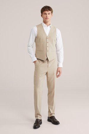 Hombre joven vestido con chaleco y pantalones beige, camisa blanca de vestir y mocasines marrón oscuro, posando frente a un fondo liso.