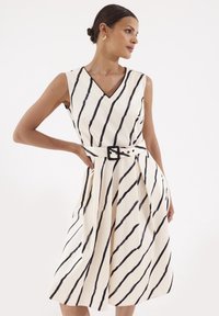 Sukienka na ramiączkach z dekoltem w serek w kolorze kremowym z czarnymi pionowymi paskami, z paskiem w talii i zwiewnym dołem. Tkanina ma gładką fakturę.