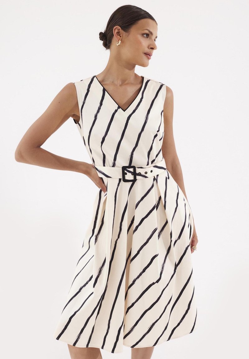 Sukienka na ramiączkach z dekoltem w serek w kolorze kremowym z czarnymi pionowymi paskami, z paskiem w talii i zwiewnym dołem. Tkanina ma gładką fakturę.