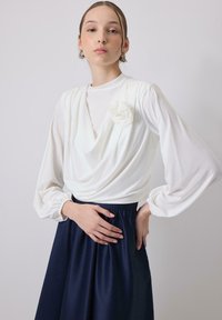 Blusa bianca drappeggiata con maniche lunghe, caratterizzata da un dettaglio di fiore in tessuto. Abbinata a una gonna fluida blu navy, enfatizzando morbide trame e contrasto.