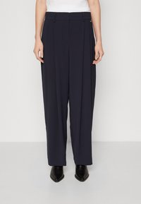 Löysät housut tummansinisestä kankaasta, joissa on etumaiset laskokset, suora istuvuus ja sileä rakenne. Täydentää hienovarainen metallikoriste.