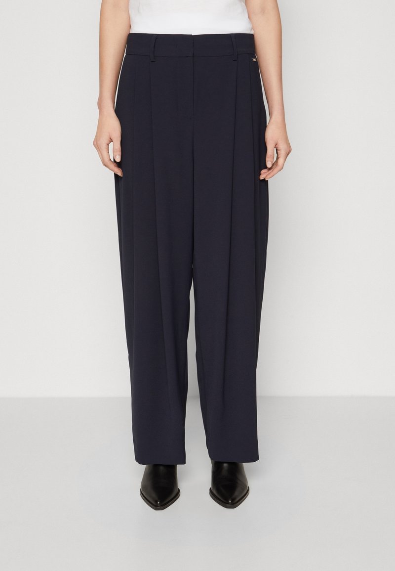 Löysät housut tummansinisestä kankaasta, joissa on etumaiset laskokset, suora istuvuus ja sileä rakenne. Täydentää hienovarainen metallikoriste.