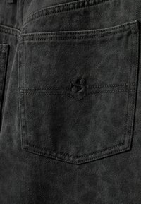 Črna denimska tkanina, ki prikazuje zadnji žep z značilnim vezenim logotipom in dvakrat šitimi šivi za trajnost.