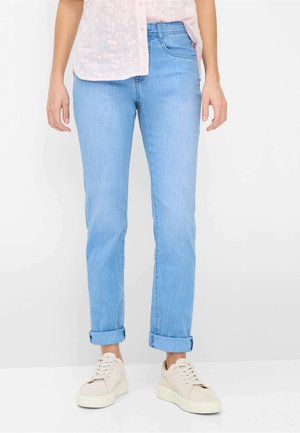 Kvinde iført lyseblå op rullede jeans, lyserød blomstret bluse og beige snørestøvler med platform, stående på hvid baggrund.