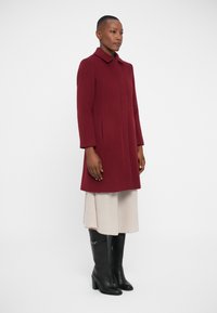 Burgunderfarbener Wollmantel mit Kragen, langen Ärmeln und seitlichen Taschen, kombiniert mit einem hellen Rock und schwarzen kniehohen Stiefeln. Glatte Textur.