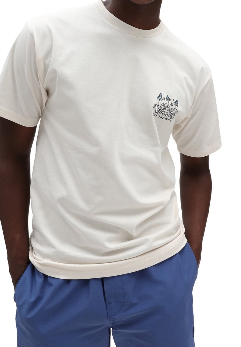 Homme portant un t-shirt Vans crème avec un logo graphique sur la poitrine et un pantalon bleu, mains dans les poches, cadré au niveau du cou et des hanches.