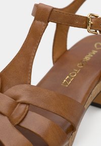 Καφέ δερμάτινο σανδάλι με ρυθμιζόμενο χρυσό αγκράφα, πλεκτές λωρίδες και χαμηλό ξύλινο τακούνι, με χρυσό κείμενο στην εσωτερική σόλα.