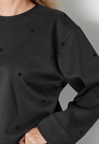 Maglione nero con una vestibilità comoda. Realizzato in tessuto texturizzato, con punti neri in rilievo e maniche ampie. Collo rotondo e leggero lucido.