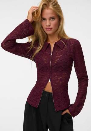 Femme blonde portant un top en dentelle bordeaux moulant à manches longues avec une fermeture éclair frontale, associé à un pantalon noir taille haute.