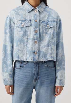 Jasnoniebieska kurtka dżinsowa z białym kwiatowym wzorem, zapinana na guziki, z klapami na kieszeniach na piersi, noszona z jeansami o średnim odcieniu.