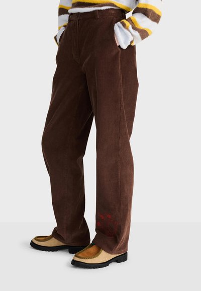 Personne portant un pantalon en velours côtelé marron avec une broderie florale rouge près de l'ourlet, des chaussures moutarde et beige, et un pull rayé avec des accents jaunes.