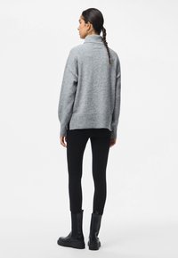 Pilkas didelio dydžio megztinis su aukštu apykakle, gilioje apdailoje ir rankogaliais, derinamas su juodomis leginsais ir juodais kulkšnies batais. Lygus fonas.